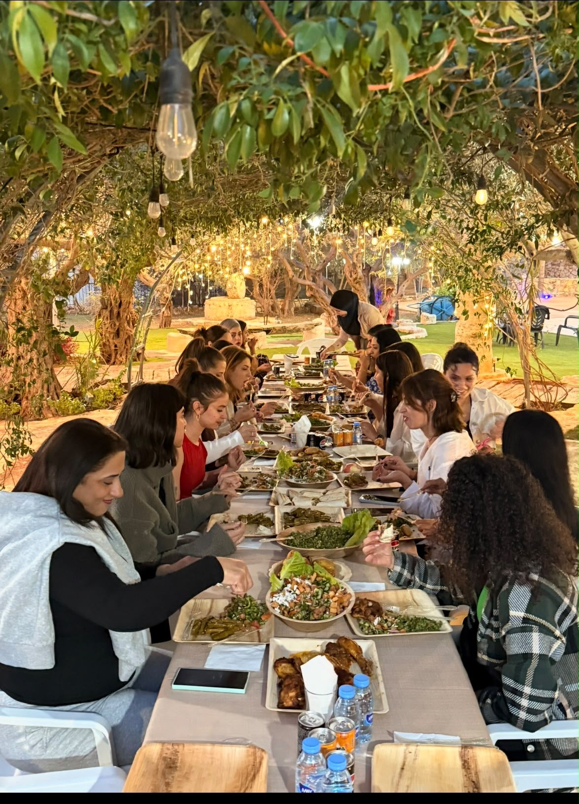 Women sharing a long communal lunch under trees and warm lights